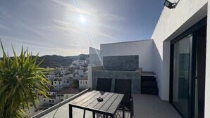 Μια ταράτσα με τραπέζι και καρέκλες, ένα φυτό σε γλάστρα και θέα προς μια πόλη στο βάθος.