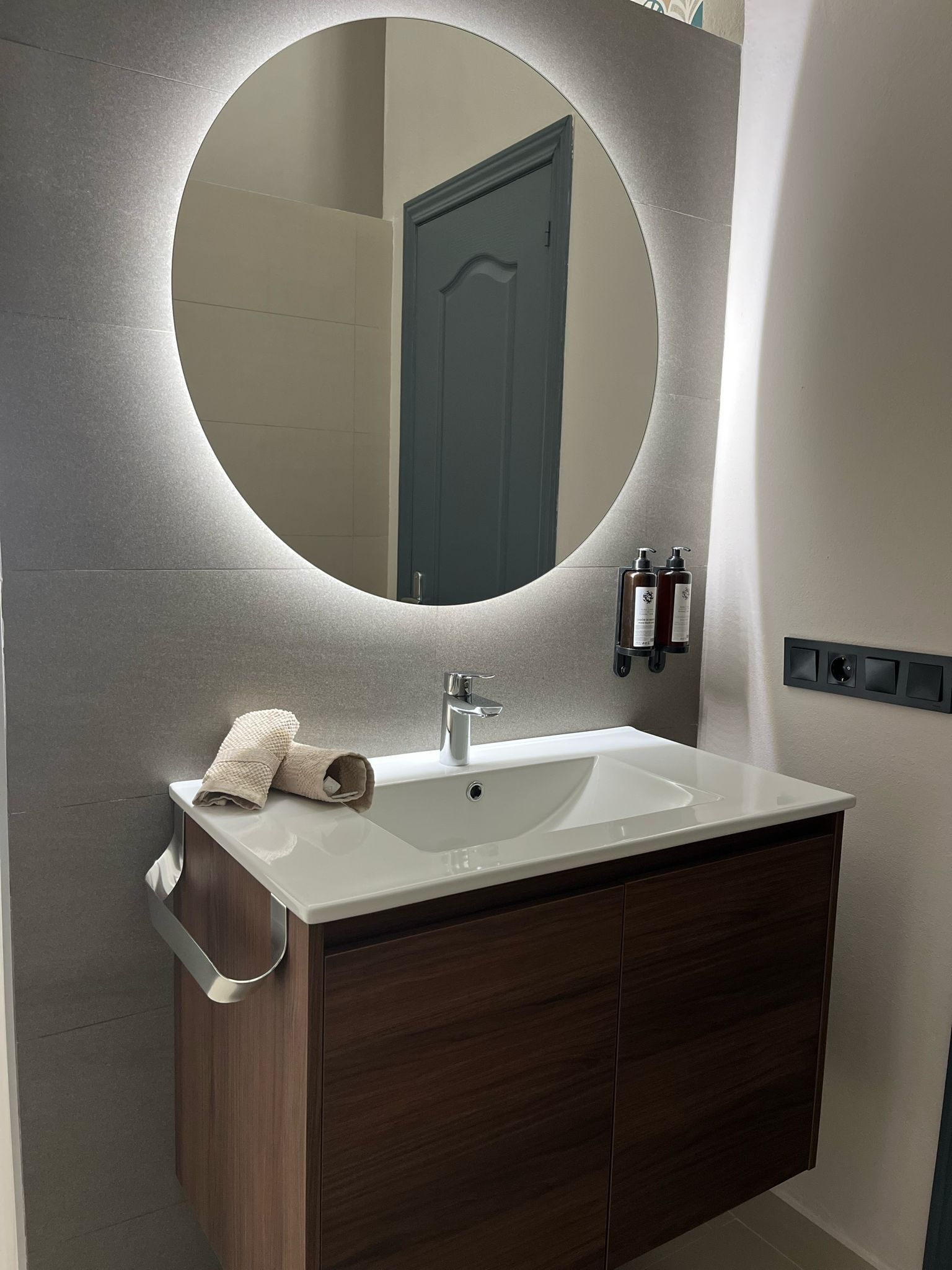 A bathroom with a round mirror, a white sink, a towel, and a door in the background.