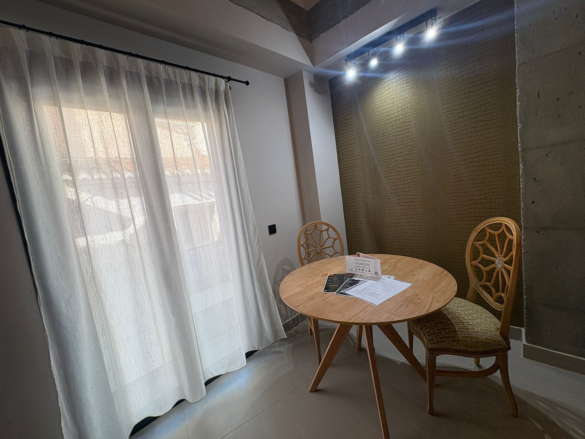 Una habitación con una mesa redonda de madera, dos sillas de mimbre, una cortina blanca y una pared con textura.
