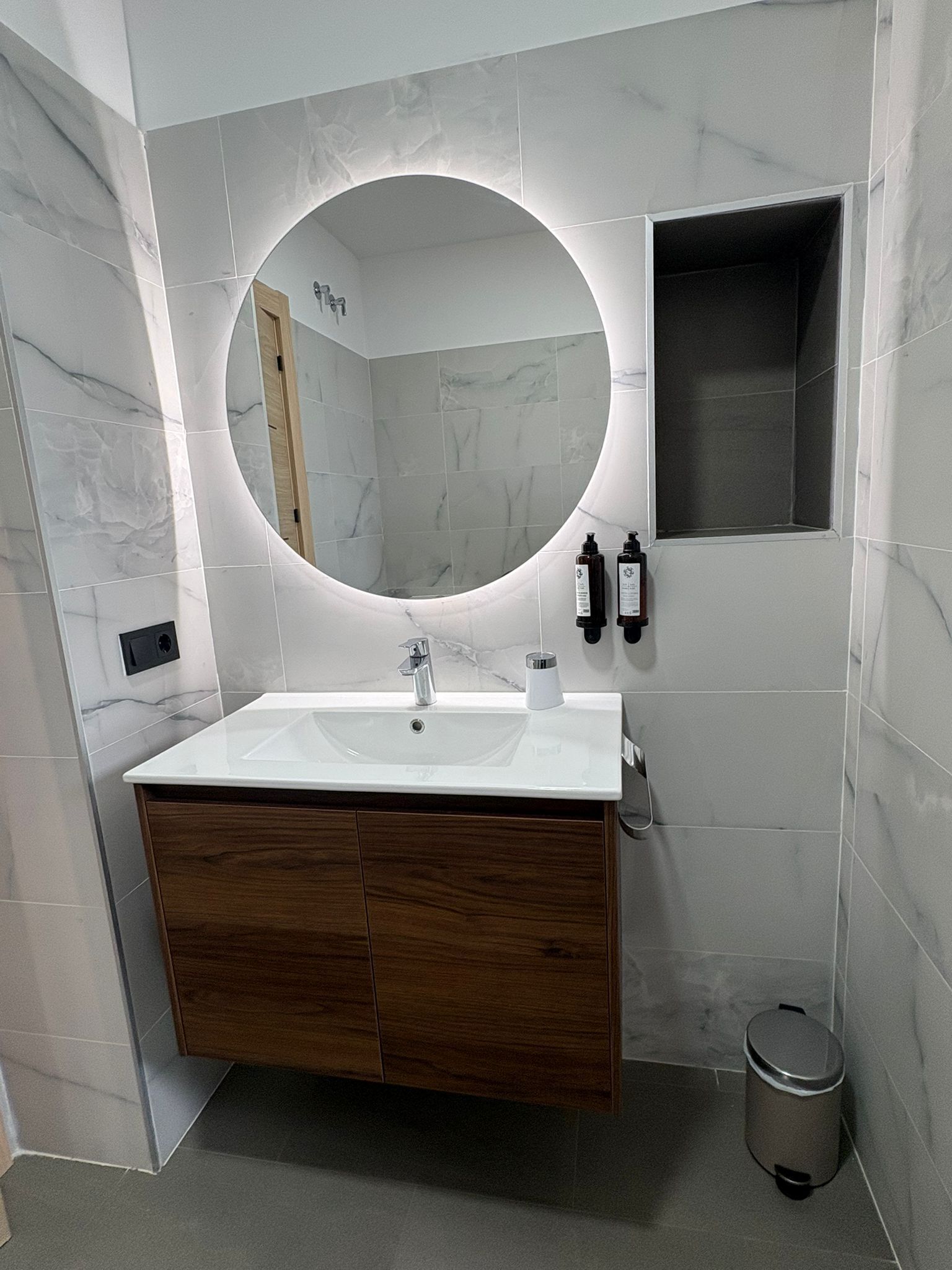 A bathroom with a white marble sink, wooden vanity, and a round mirror with a light.