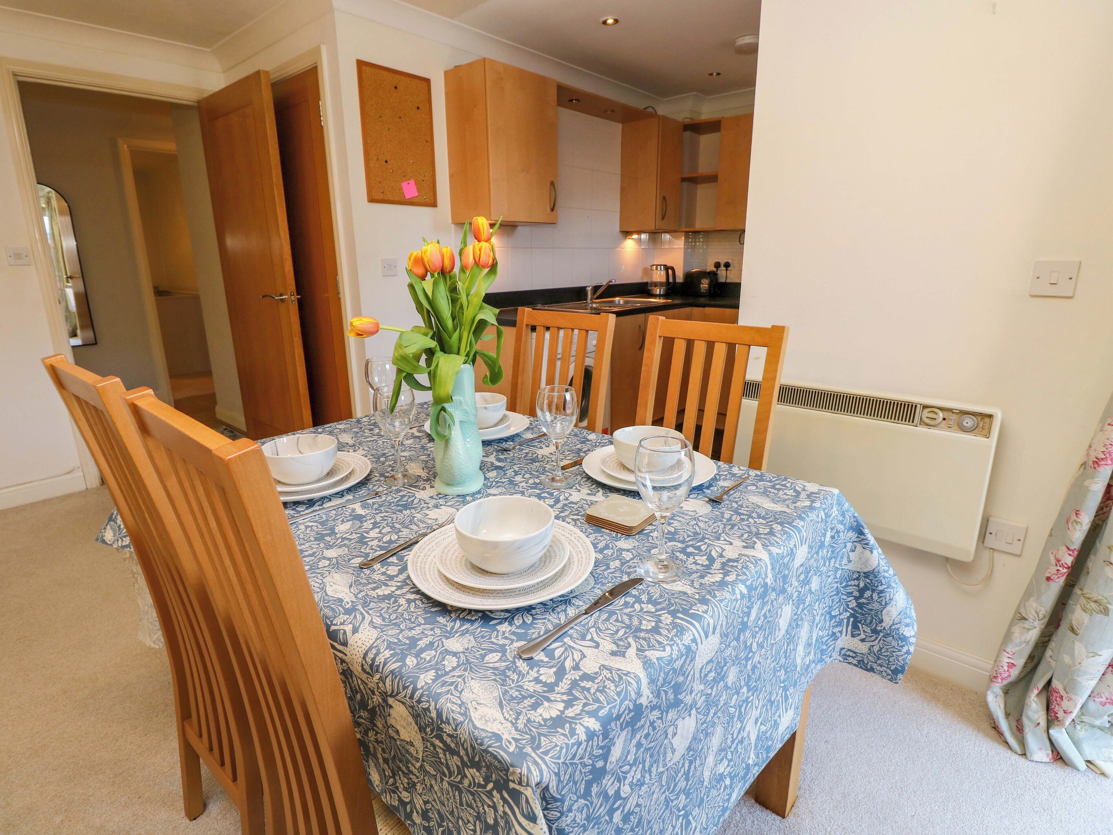 A dining area with a table set for four, surrounded by wooden chairs. A vase with flowers is on the table, and there are kitchen cabinets in the background.
