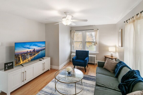 A living room with a flat-screen TV, a ceiling fan, a blue armchair, a green sofa, and a small table with a lamp.