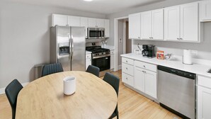 A modern kitchen with white cabinets, stainless steel appliances, and a wooden dining table. - Highwood Haven- 4 Bedroom Steps to Train station! (Highwood)