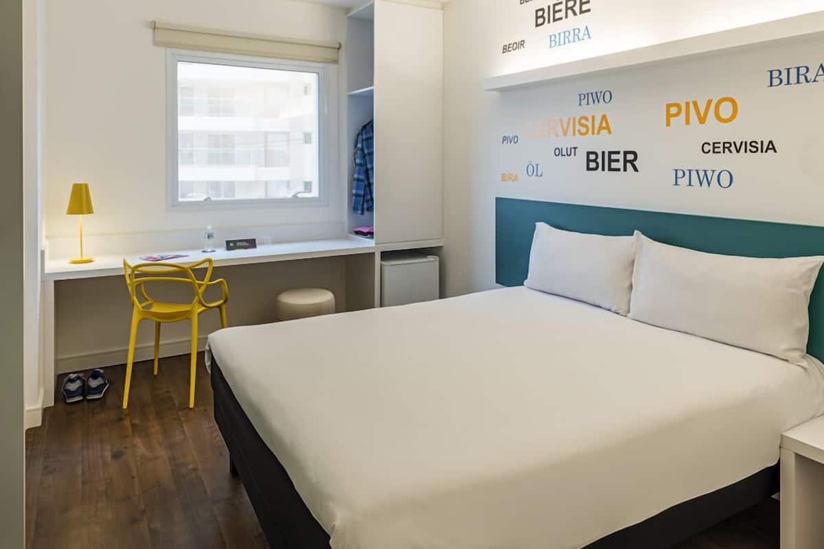 A hotel room with a bed, a desk, a chair, a window, and a wall with words in different languages.