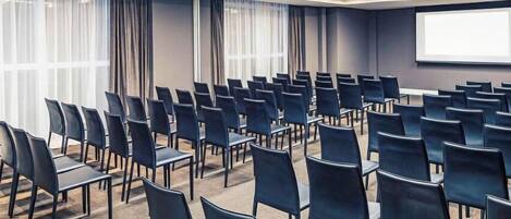 Un salón de conferencias con filas de sillas negras, una pantalla grande y cortinas en las ventanas.