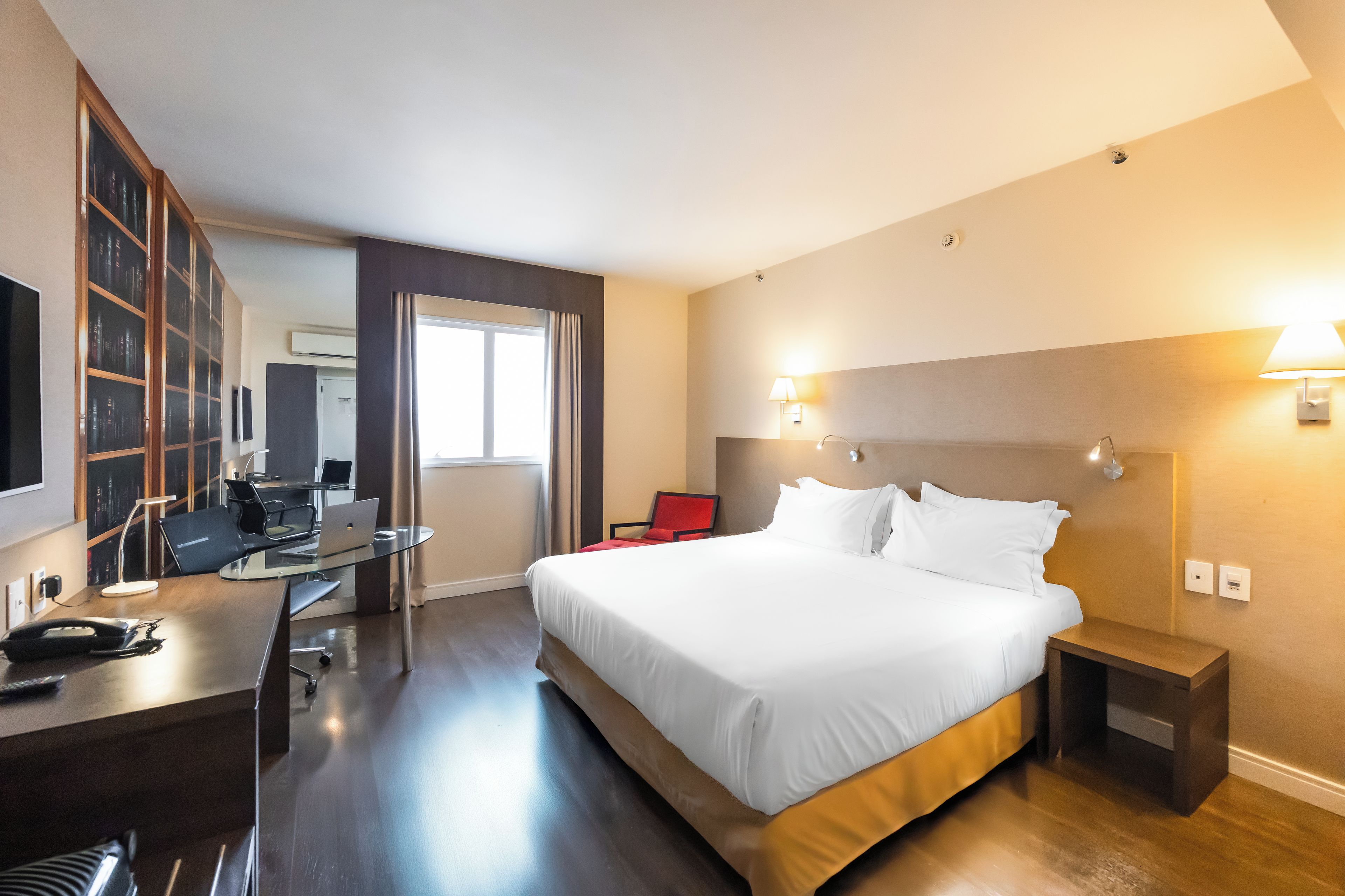 Una habitación de hotel con una cama, un escritorio con una computadora portátil, una silla, una estantería y un televisor.