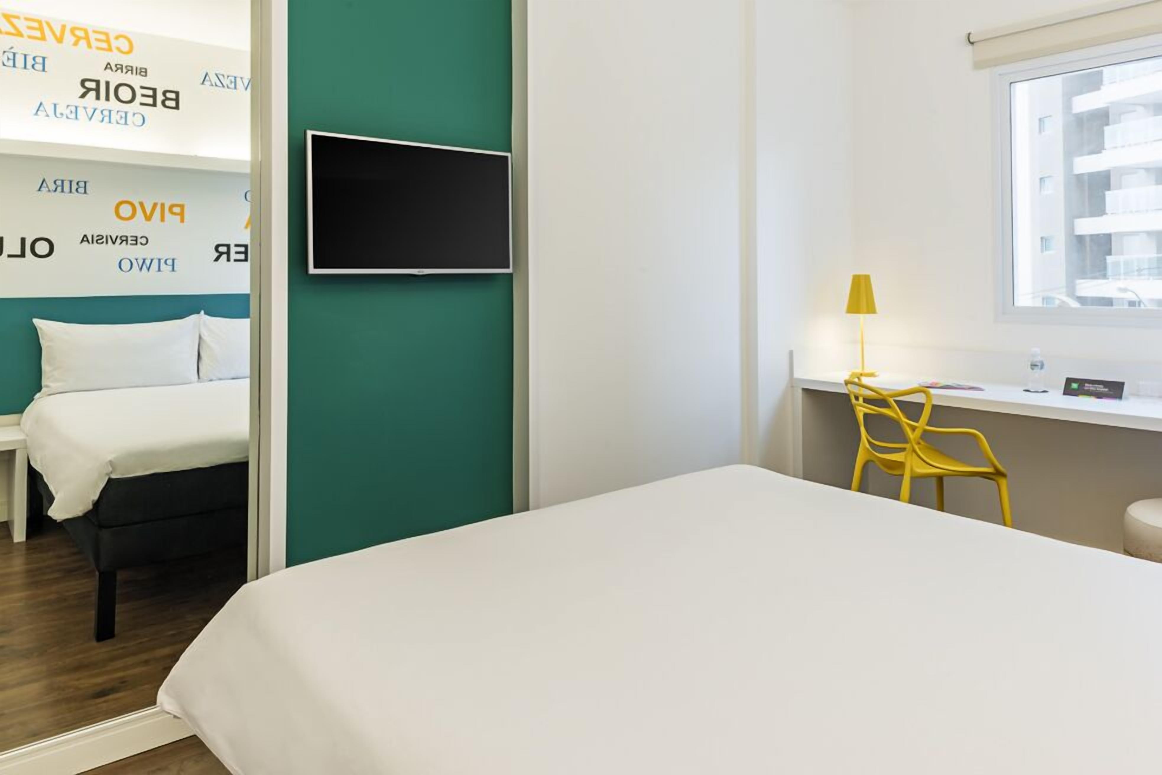 Habitación de hotel con cama, escritorio con silla, televisión y ventana con vista a edificios.