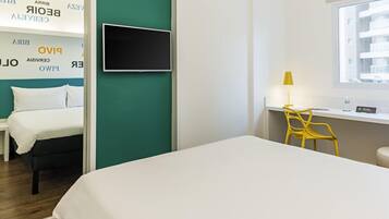 Habitación de hotel con cama, escritorio con silla, televisión y ventana con vista a edificios.