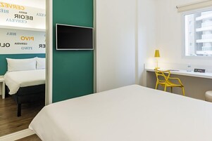 Habitación de hotel con cama, escritorio con silla, televisión y ventana con vista a edificios.