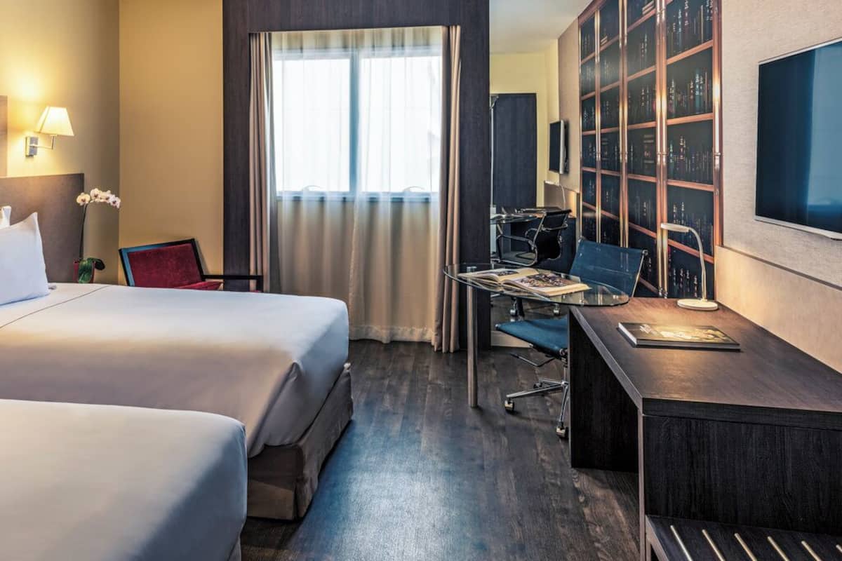 A hotel room with two beds, a desk, a chair, a TV, and a bookshelf.