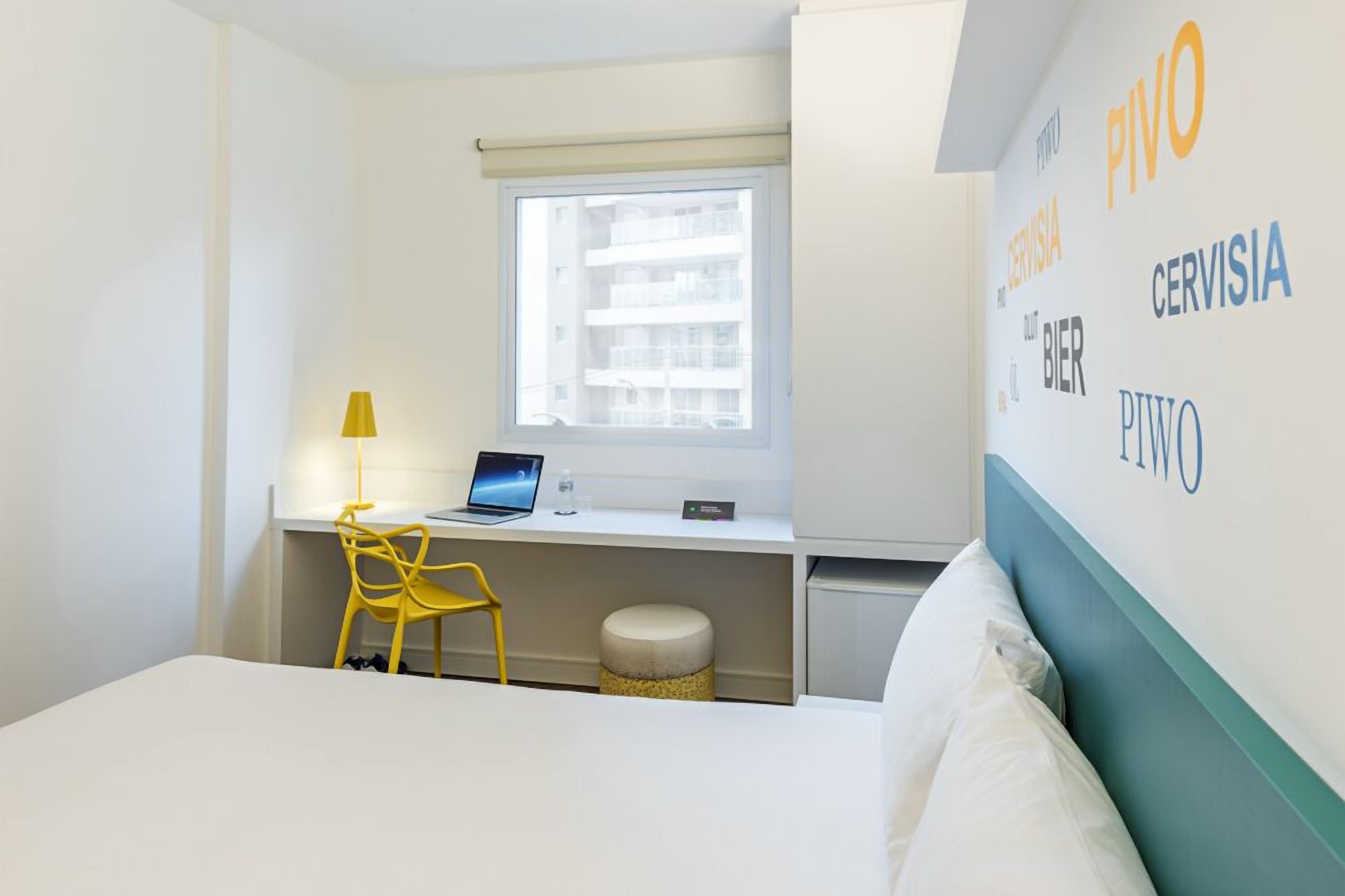 A hotel room with a bed, a desk with a laptop, a chair, a window, and a wall with text.