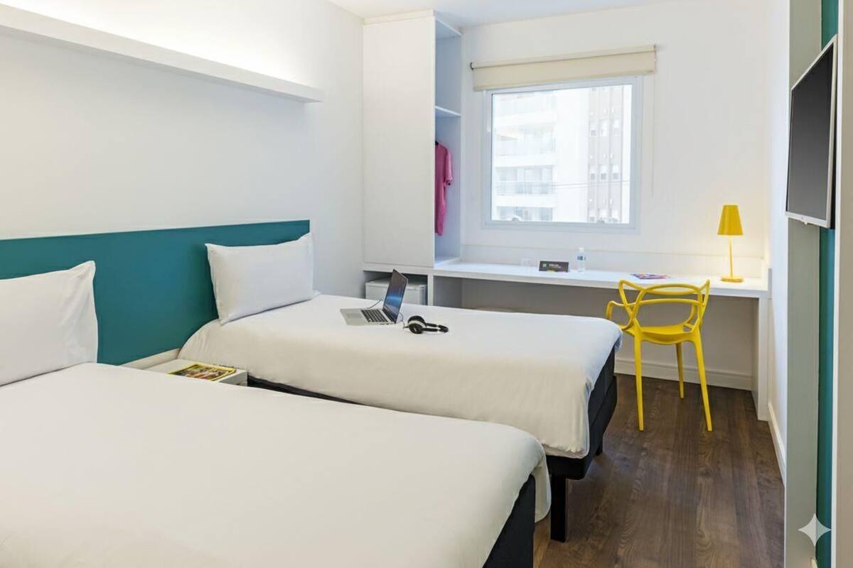 A hotel room with two beds, a desk with a laptop, a chair, and a window with a view of buildings.