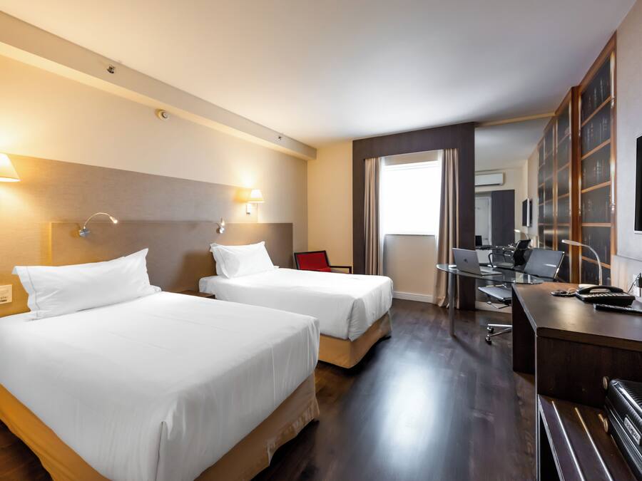 Habitación de hotel con dos camas, un escritorio con una computadora portátil y un televisor grande en la pared.