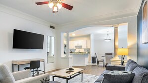 A modern living room with a flat-screen TV, a ceiling fan, and a dining area with a kitchen in the background.