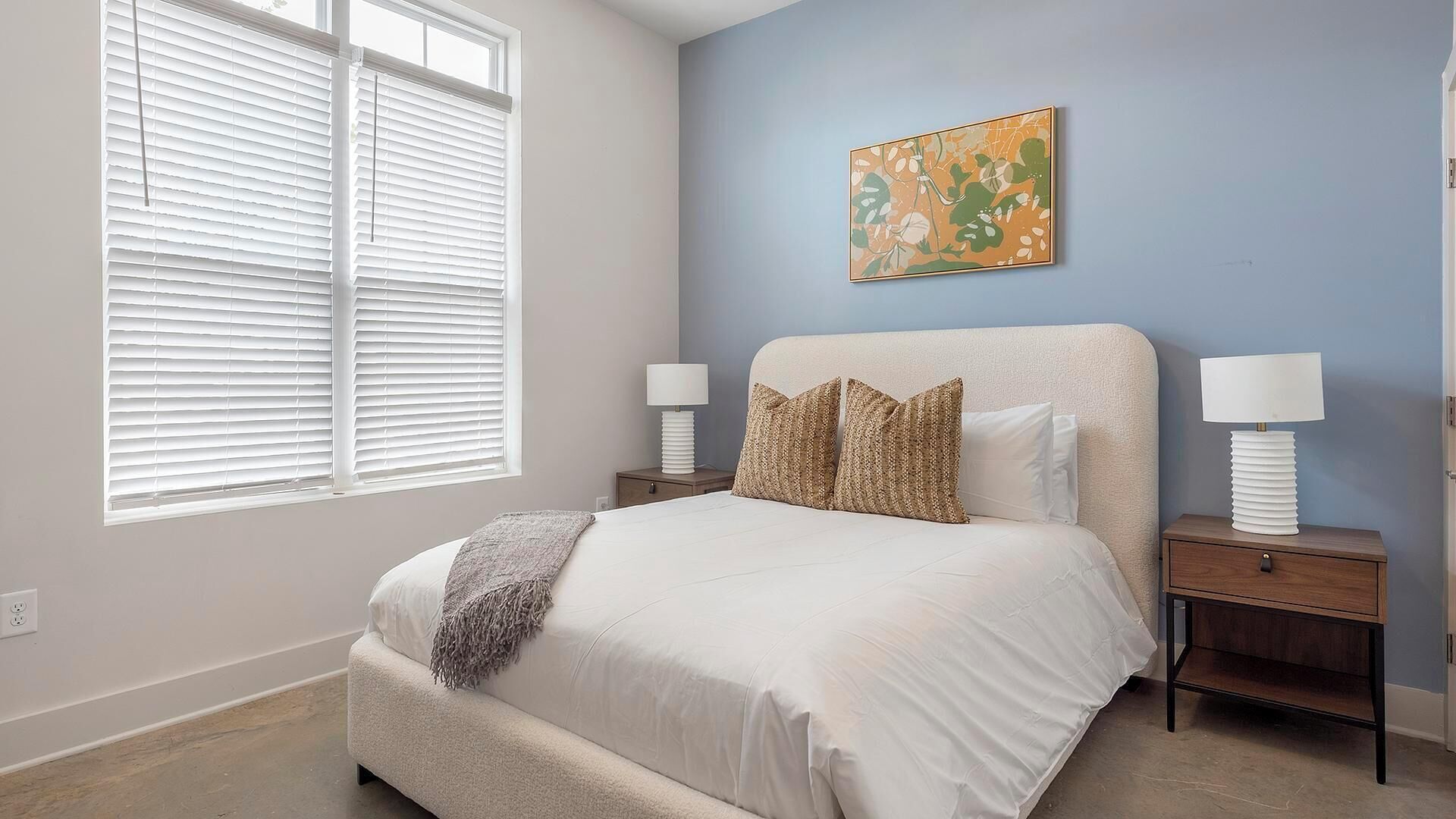 A bedroom with a large bed, two bedside tables with lamps, a window with blinds, and a wall with a piece of abstract art.