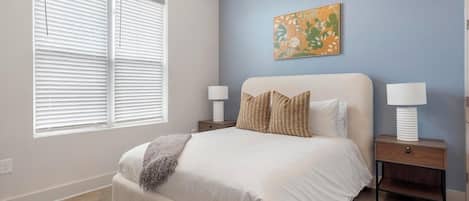 A bedroom with a large bed, two bedside tables with lamps, a window with blinds, and a wall with a piece of abstract art.