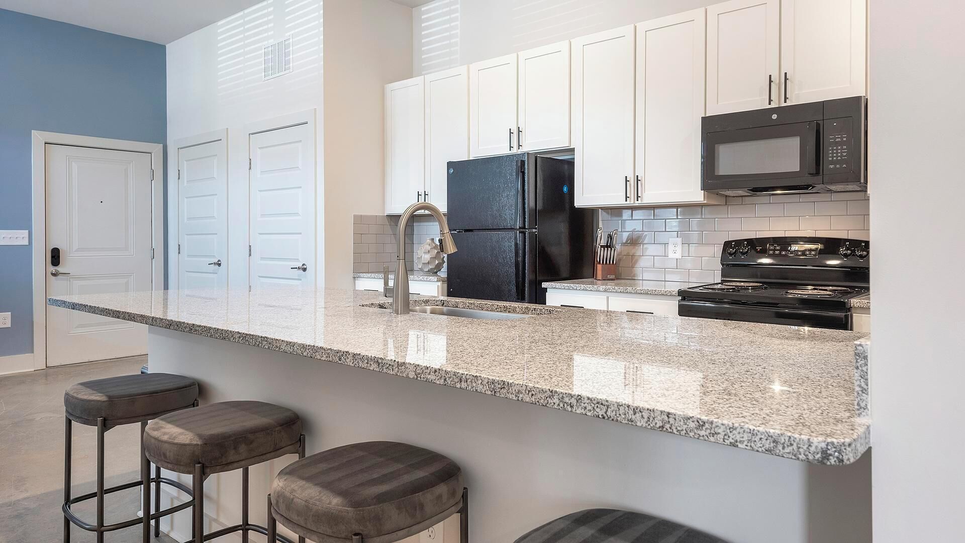 A modern kitchen with white cabinets, a black refrigerator, a microwave, and a stove. There are three bar stools and a window with blinds.