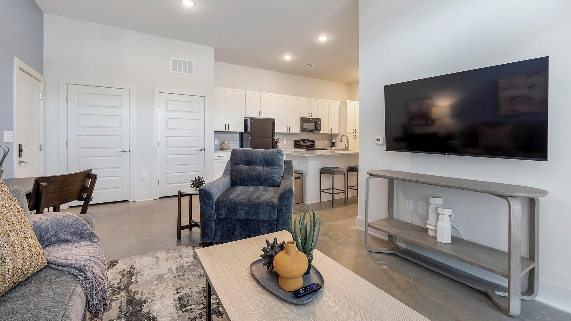 A modern living room with a sofa, a coffee table with decorative items, a TV mounted on the wall, and a kitchen area with white cabinets and appliances.
