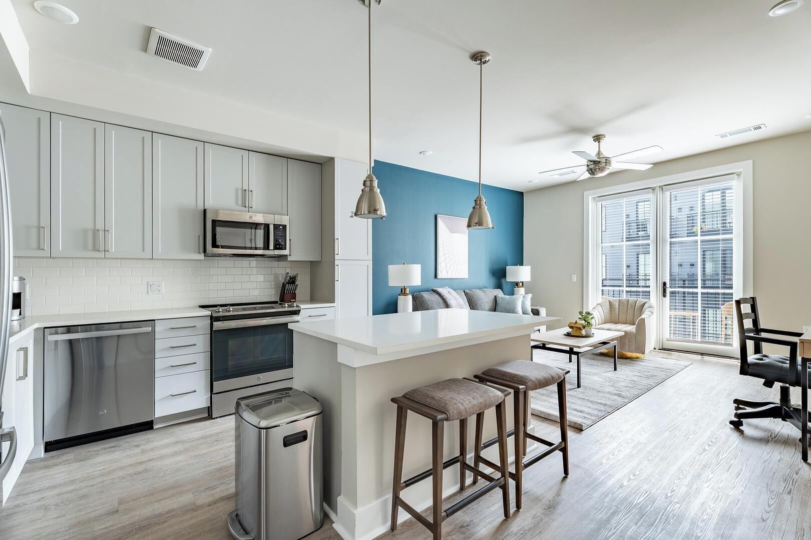 A modern kitchen with white cabinets, a microwave, and a stainless steel dishwasher. There is a bar area with stools and a seating area with a sofa and chairs.