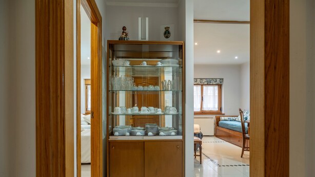 Una habitación con un armario de madera que contiene cristalería y vajilla, una cama con colcha azul y una ventana con cortinas.