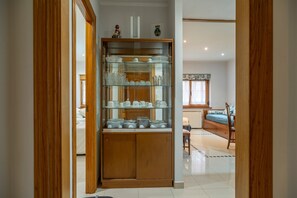 A room with a wooden cabinet containing glassware and dishes, a bed with a blue bedspread, and a window with curtains. - Apartment 'El Fondón 1' in Cangas de Onís – Central, Pet-Friendly, Step-Free Access (Cangas de Onís)