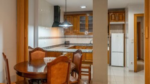 Un comedor con mesa y sillas de madera, una cocina con armarios de madera y un refrigerador blanco, y un pasillo con puertas de madera.