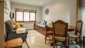 Una sala de estar acogedora con un sofá, una mesa de comedor con sillas, un televisor montado en la pared y un ventanal con cortinas.