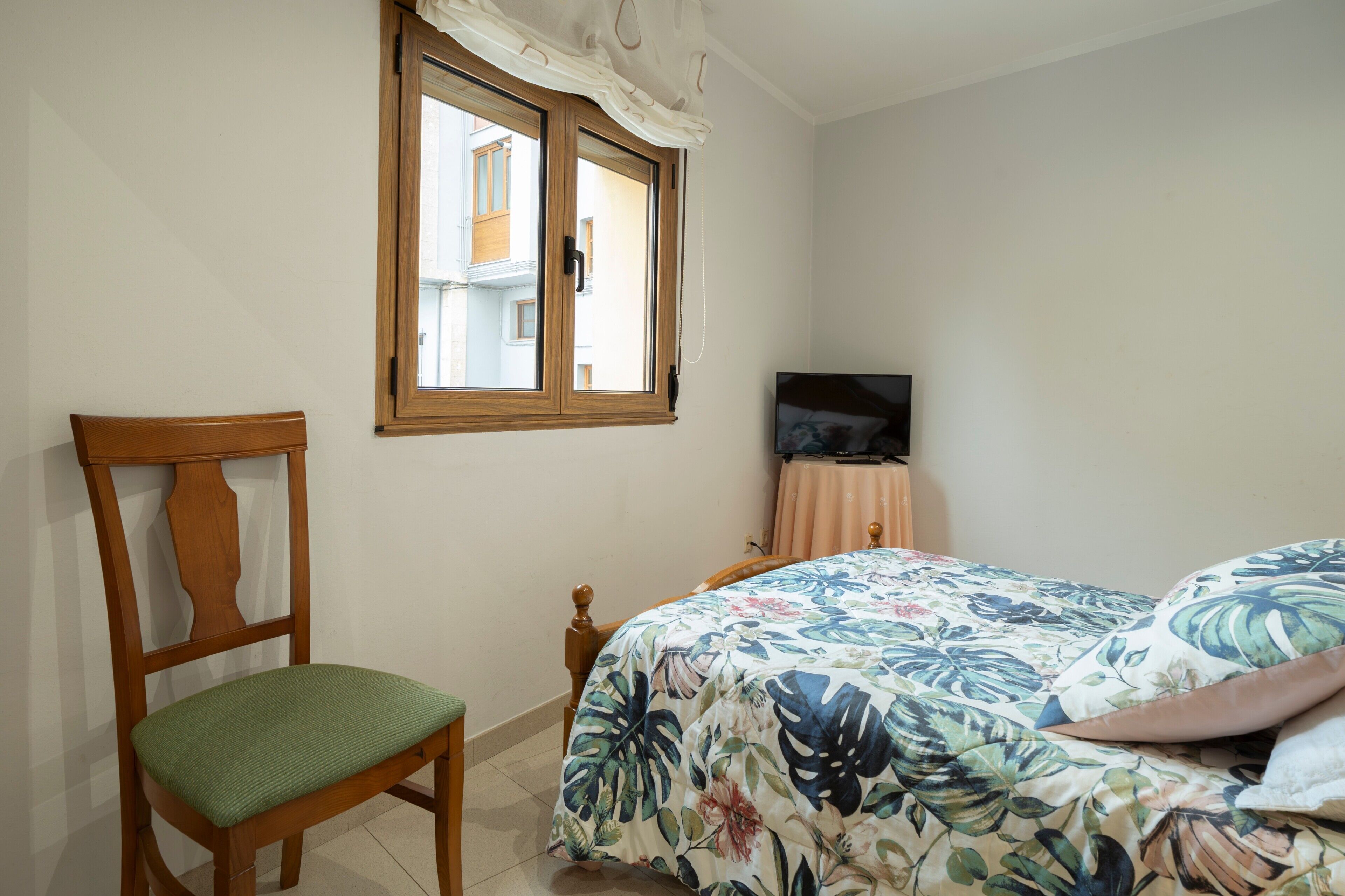 A bedroom with a bed, a chair, a television on a stand, and a window with curtains.