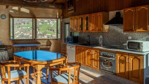 A kitchen with wooden cabinets, a granite countertop, a dining table with chairs, and a view of a green landscape through large windows. - Apartment 'Casa Geli 2' in La Robellada, Onís – Up to 9 Guests, 4 Bedrooms, 2 Bathrooms (La Robellada)