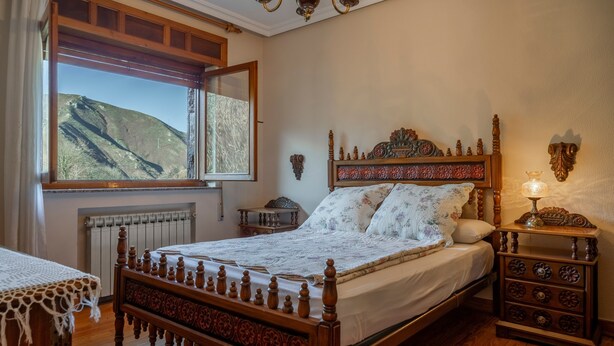 A bedroom with a wooden bed, a nightstand, a lamp, a window with a view of a mountain, and a radiator. - Apartment 'Casa Geli 2' in La Robellada, Onís – Up to 9 Guests, 4 Bedrooms, 2 Bathrooms (La Robellada)