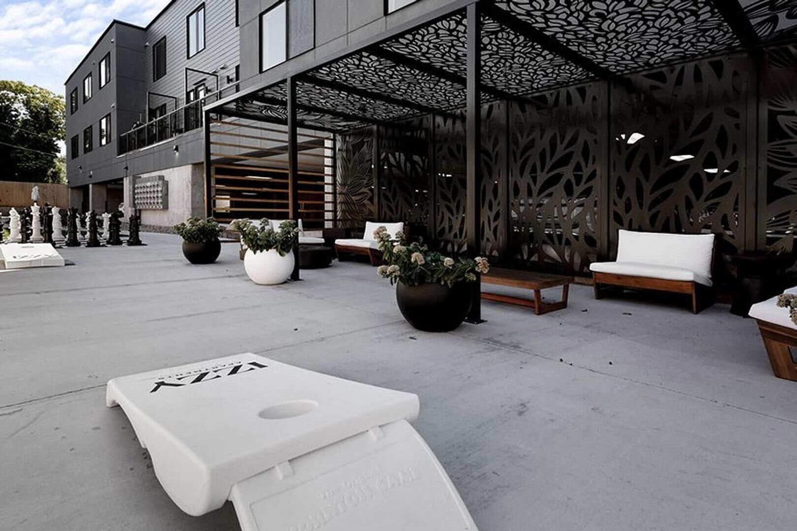 An outdoor seating area with modern furniture and potted plants, a covered section with intricate lattice design, and a building with a contemporary facade.