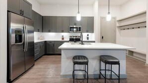 A modern kitchen with grey cabinets, stainless steel appliances, a central island with bar stools, and pendant lighting. - Landing | Beautiful 2BD, Gym, Courtyard (Austin)