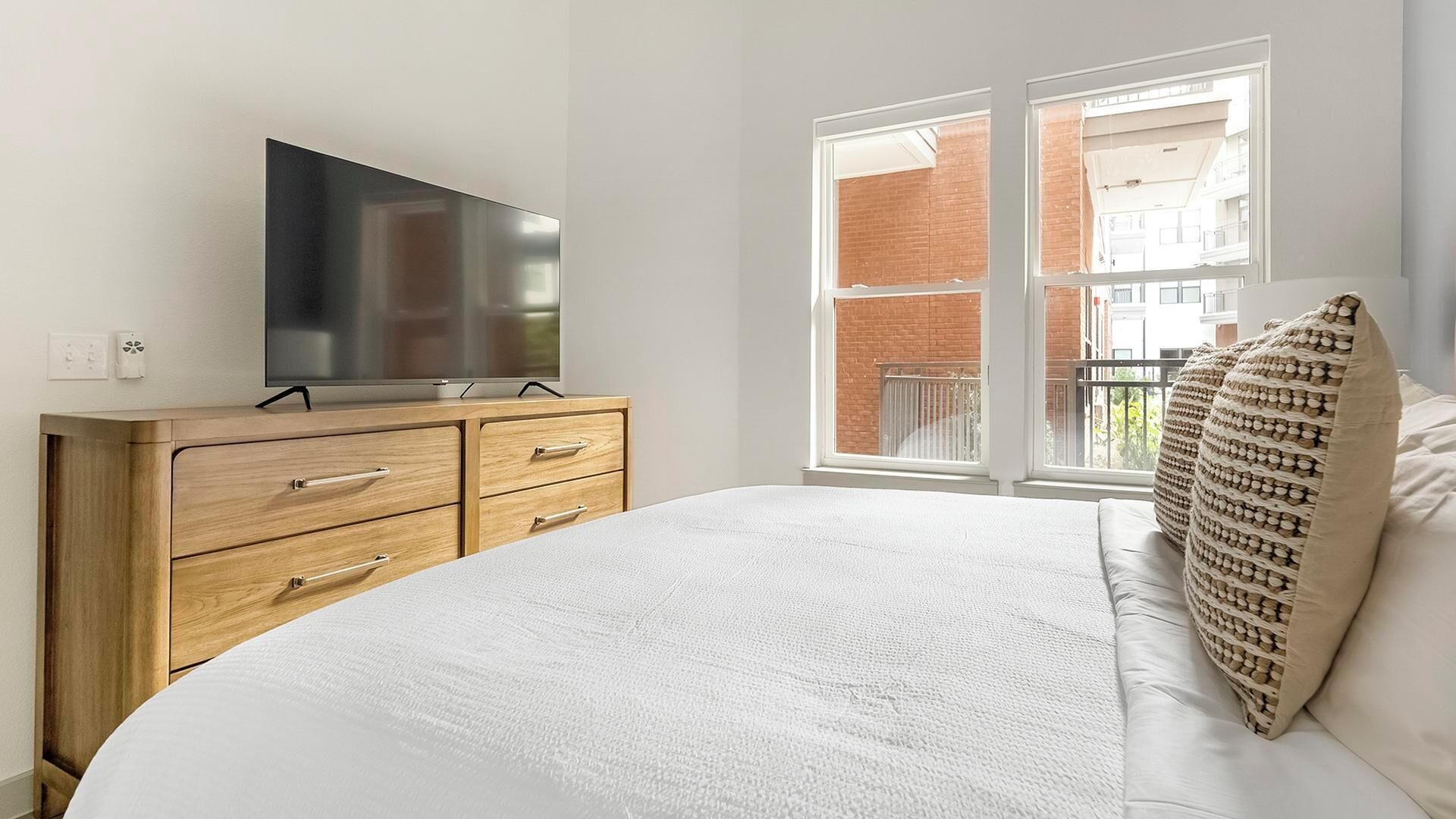 Een slaapkamer met een bed, een houten dressoir, een flatscreen televisie en een raam met uitzicht op een gebouw van baksteen.