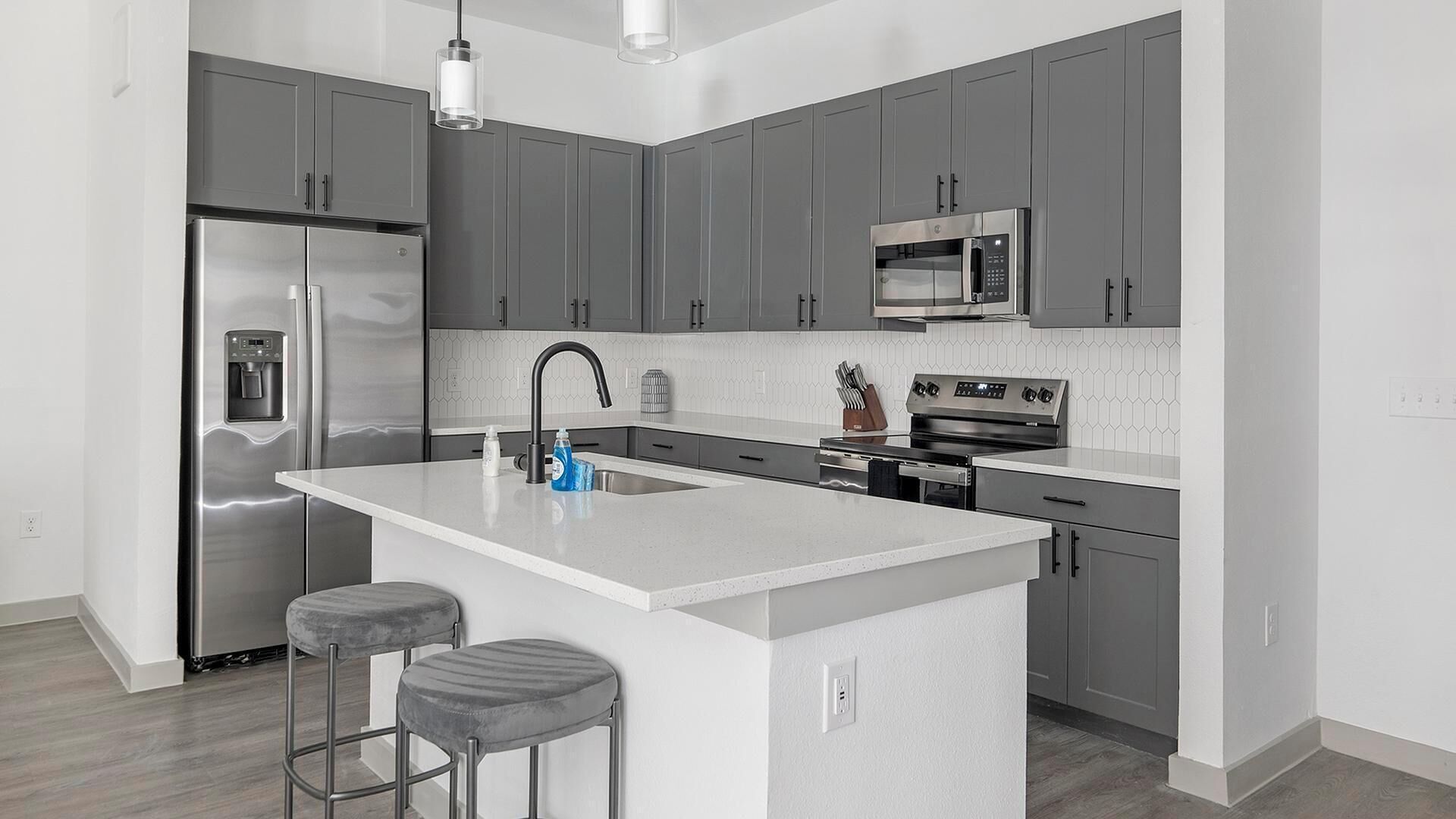 Una cocina moderna con armarios grises, electrodomésticos de acero inoxidable, una isla central con taburetes y una lámpara colgante.