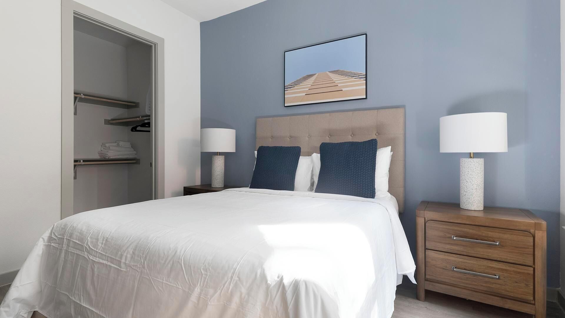 Une chambre à coucher avec un lit, deux tables de chevet avec des lampes, une armoire et un tableau encadré au mur.