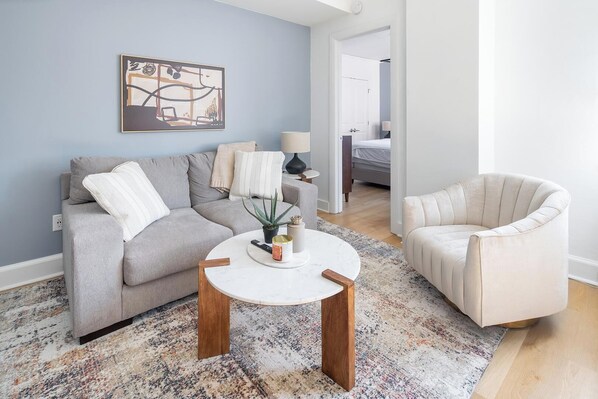 Sala de estar com sofá cinza, mesa de centro redonda e poltrona bege. Há um quadro na parede e uma planta na mesa de centro.