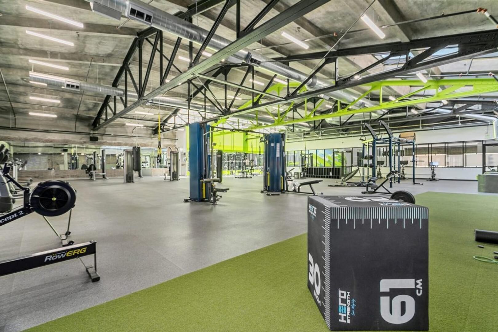 A gym with various exercise equipment, green flooring, and a high ceiling with visible structural beams.