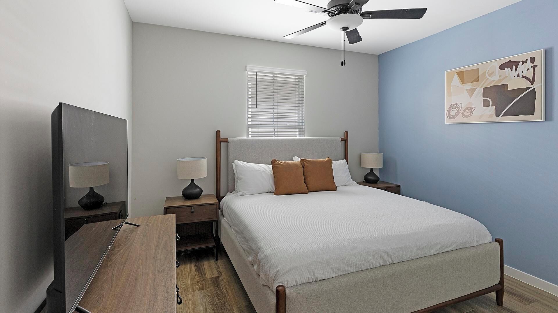 A bedroom with a bed, two bedside tables with lamps, a ceiling fan, a window with blinds, and a wall with a piece of abstract art.