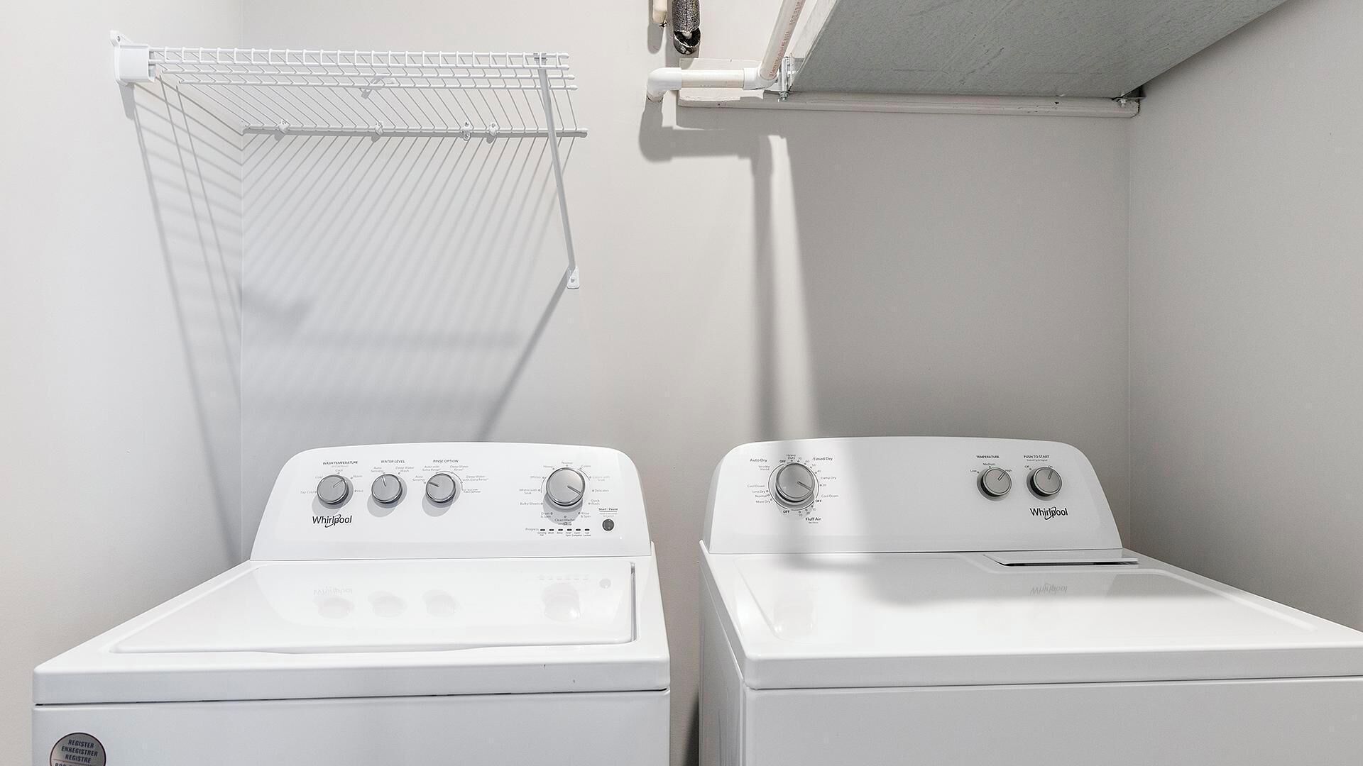 Two Whirlpool front-loading washing machines in a laundry room with a metal drying rack and a white ceiling.