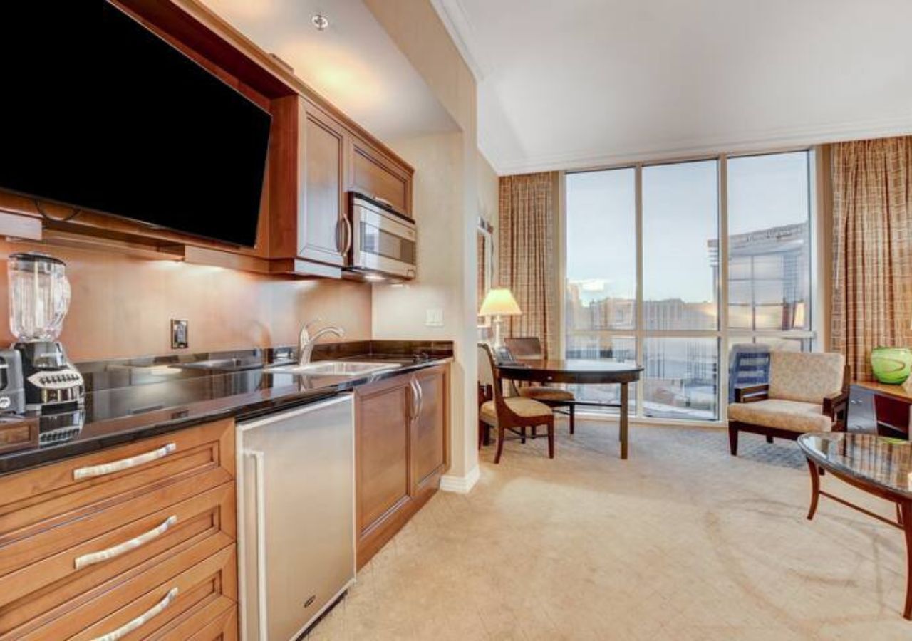 A modern hotel room with a kitchenette, a dining area, and a large window offering a city view.