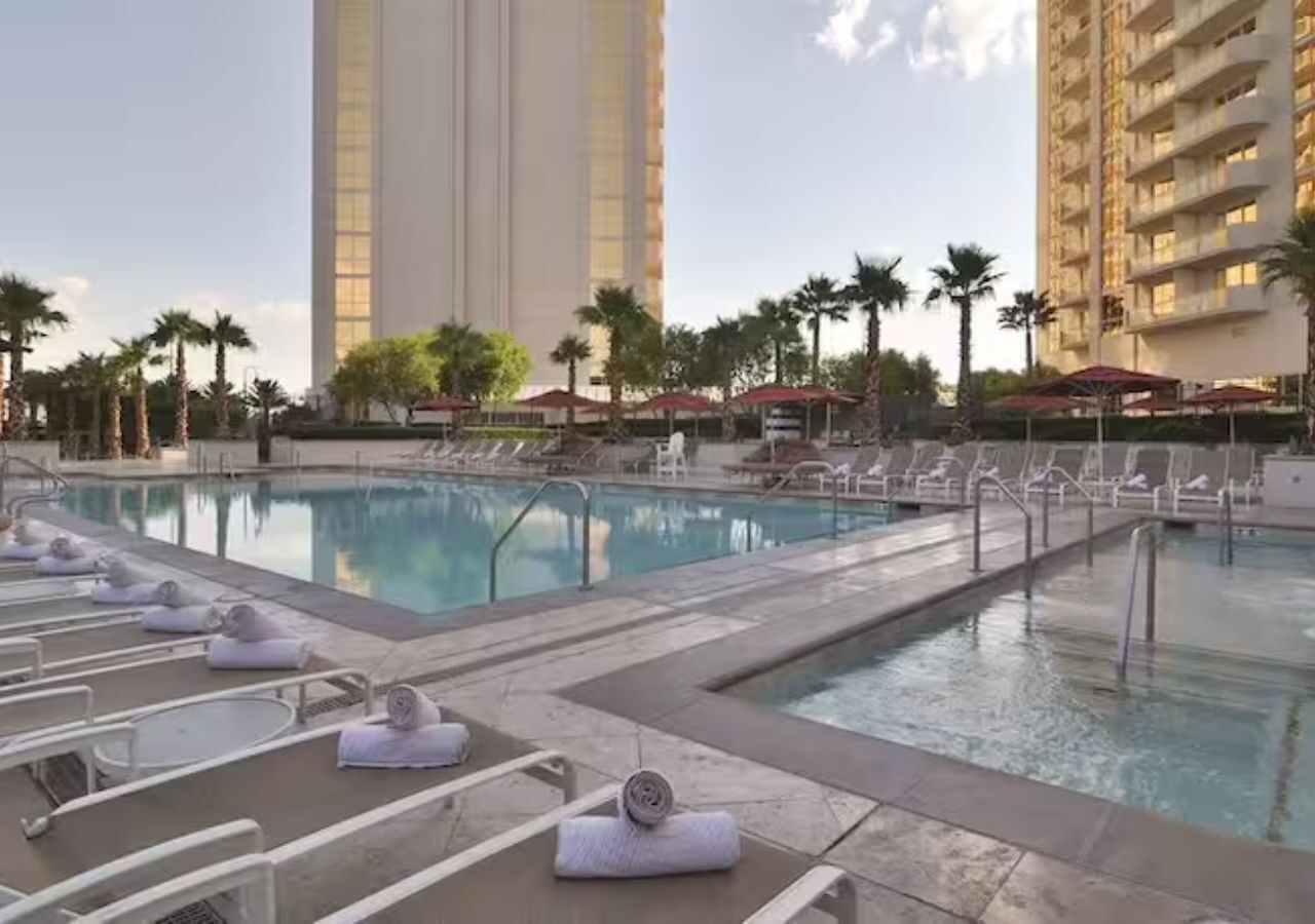 A swimming pool with lounge chairs and towels, surrounded by palm trees and tall buildings.