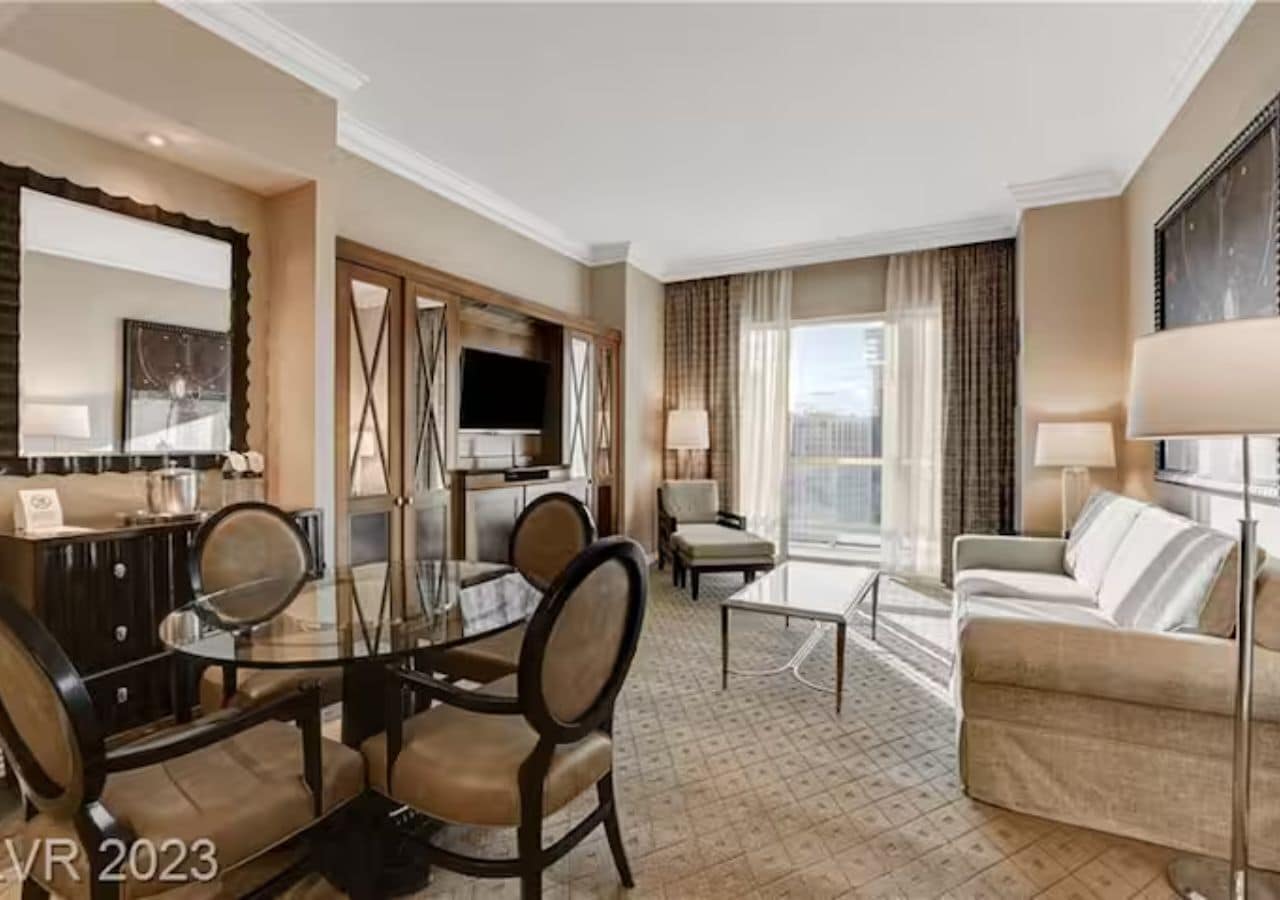 A hotel room with a dining area, a sofa, a TV, and a large window with curtains.