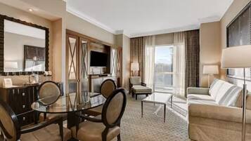 A hotel room with a dining area, a sofa, a TV, and a large window with curtains.