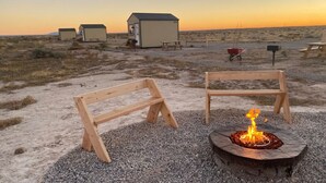 A desert campsite with wooden benches, a fire pit, and small cabins in the background.