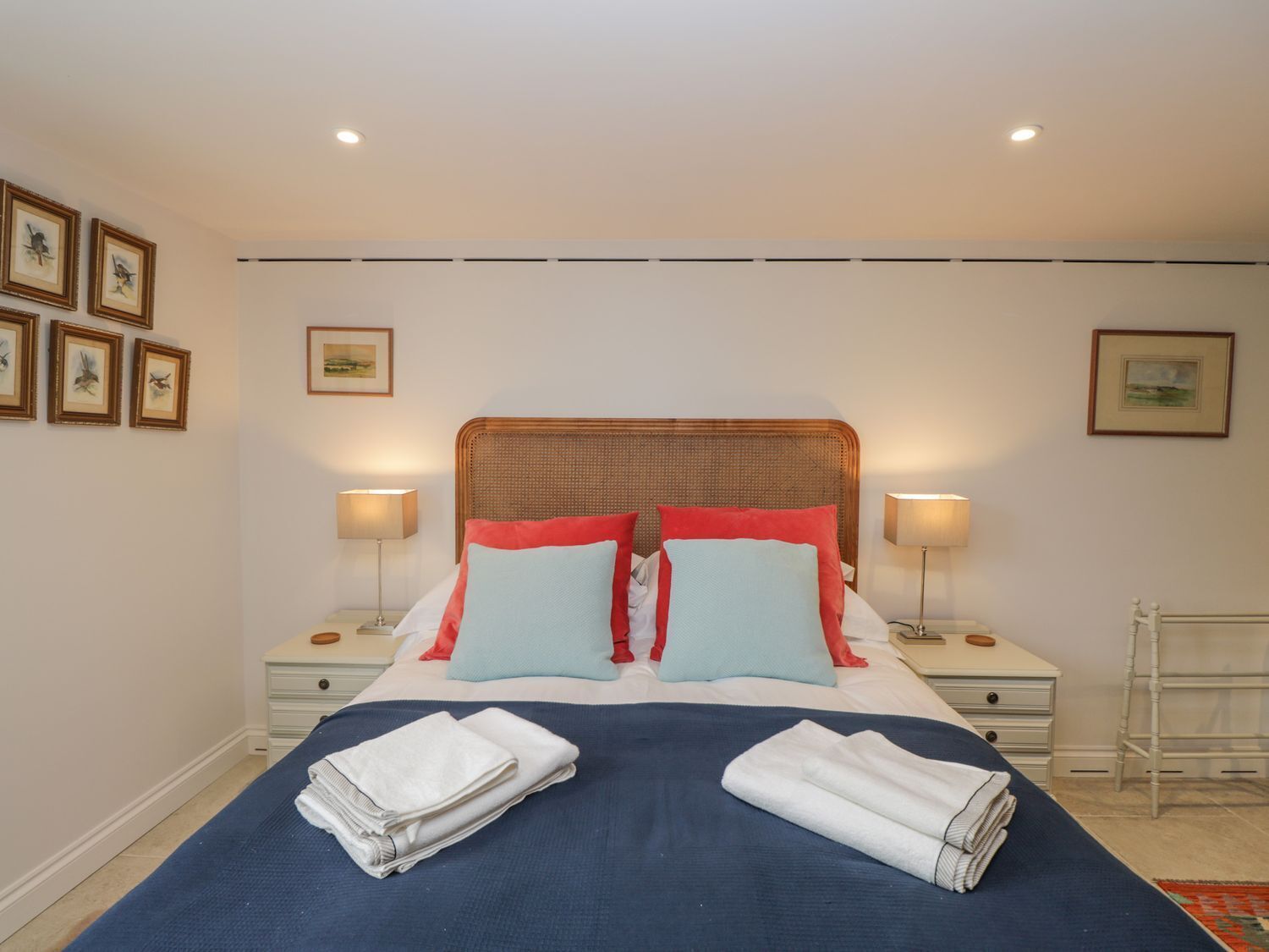 A neatly made bed with a blue bedspread, two white towels, and a wicker headboard. There are two bedside tables with lamps, a white chair, and framed pictures on the walls.