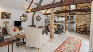Un ampio soggiorno con soffitto alto, travi in legno a vista, un caminetto e una cucina sullo sfondo.