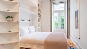 A bedroom with a bed, white shelving units, a potted plant, and a door leading to a balcony. - Porto Tiles | 2BR Duplex + Studio by LovelyStay (Porto)