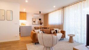Ein modernes Wohnzimmer mit einer Couch, einem Couchtisch und einem Fernseher. Im Hintergrund ist eine Küchenzeile mit einem Esstisch und Stühlen zu sehen.