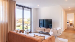Ein modernes Wohnzimmer mit Sofa, Fernseher und Blick auf einen Park durch große Fenster.