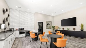 Uma cozinha moderna com armários brancos, bancada preta e backsplash. Há duas mesas redondas com cadeiras laranja. Uma televisão de tela plana grande está fixada na parede.